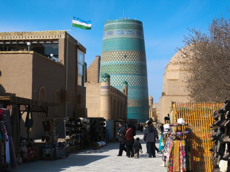 uzbekistan come pagare Khiva: 10 cose da vedere a Itchan Kala in 1 o 2 giorni (2026) Il minareto Kalta Minor di Khiva, basso e rivestito di maiolica turchese, con la bandiera dell'Uzbekistan sugli edifici di mattoni di Itchan Kala