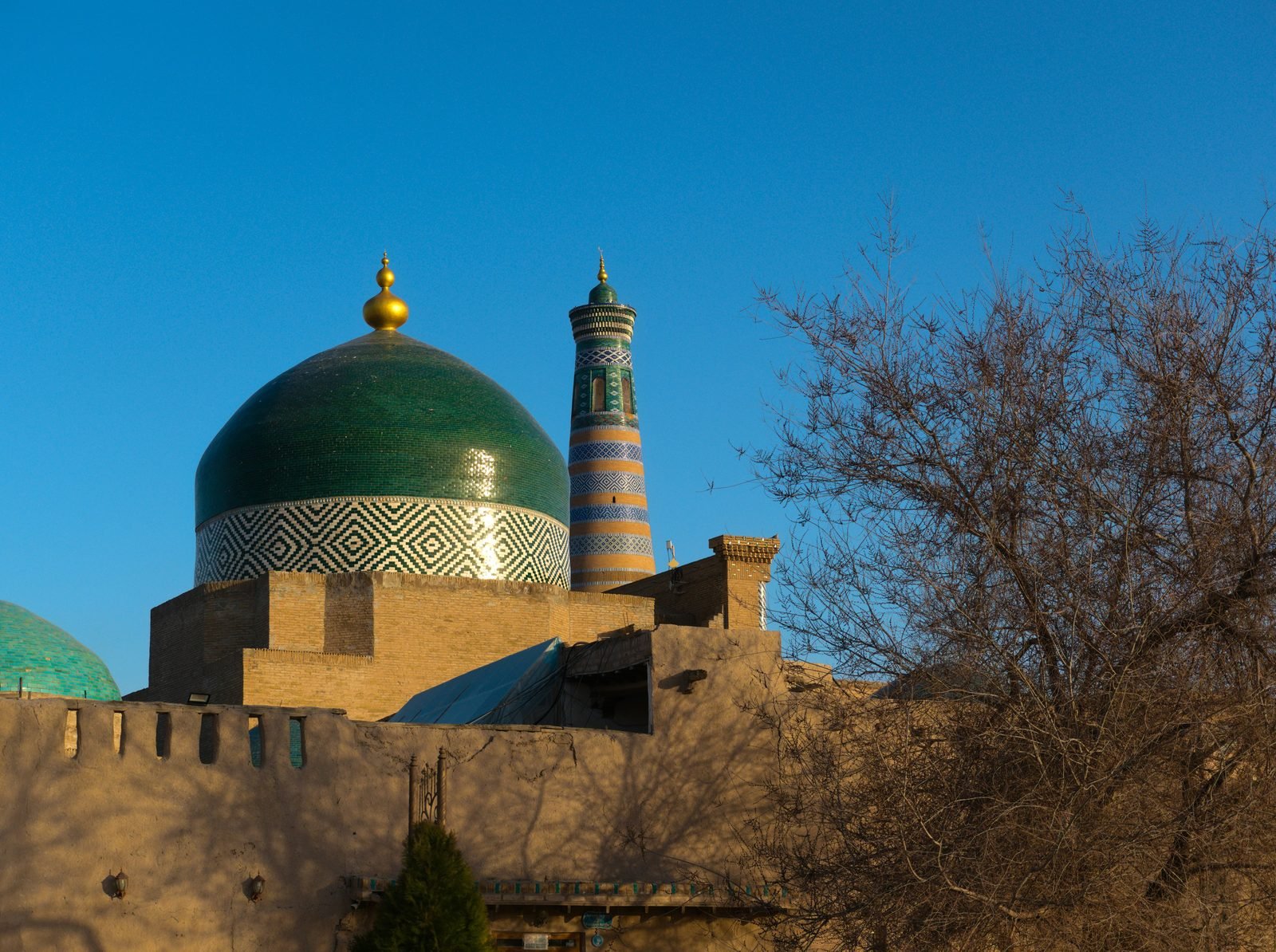khiva cosa vedere Khiva: 10 cose da vedere a Itchan Kala in 1 o 2 giorni (2026) Il minareto Islam Khoja a fasce di maiolica dietro la cupola turchese del mausoleo di Pahlavan Mahmud a Khiva