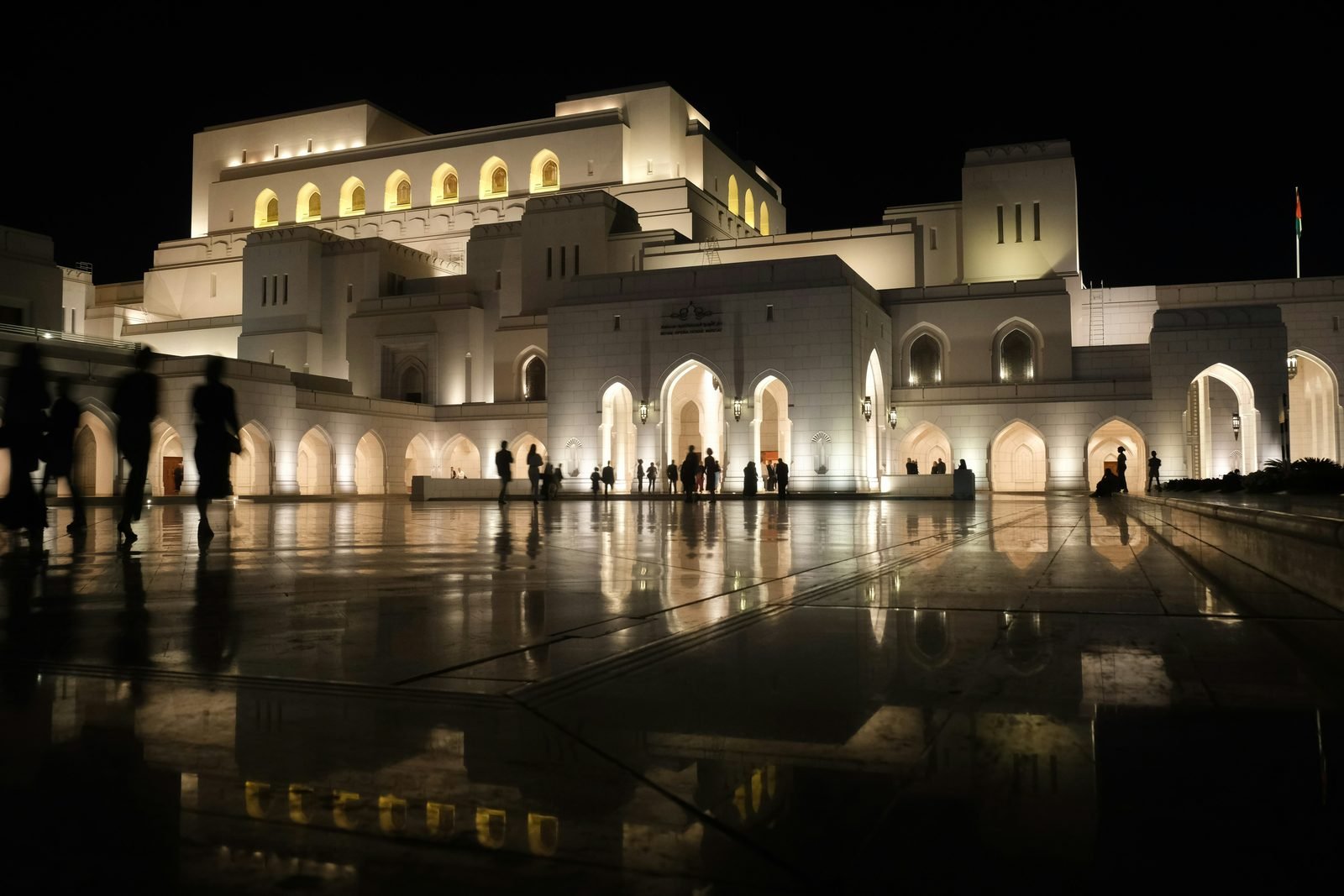 La Royal Opera House di Muscat illuminata di notte, con la facciata bianca ad archi riflessa sul selciato bagnato